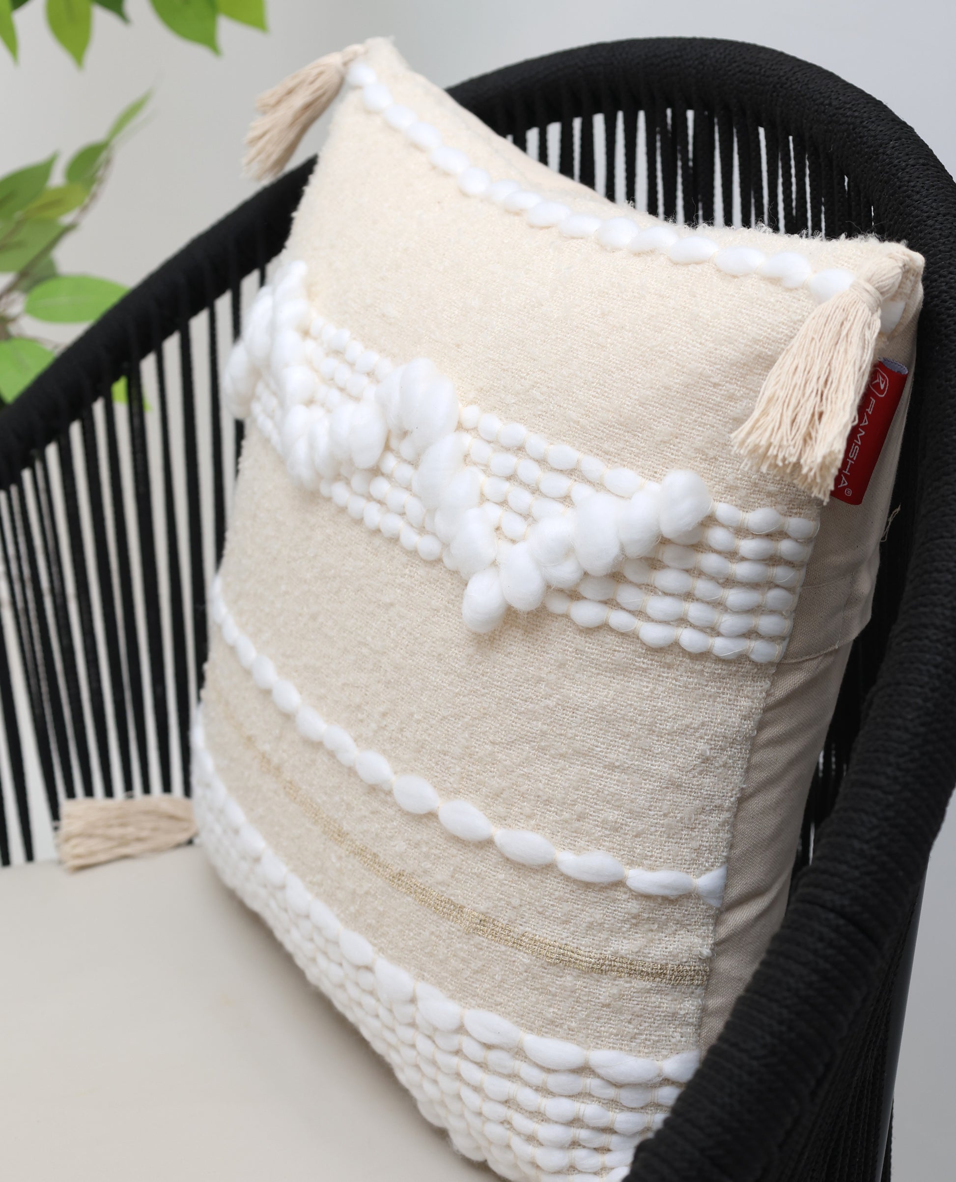 The Cream Color Home Decor Cushion - CS 313 A by Ramsha Home features premium cotton with white textured patterns and tassels, displayed on a black woven chair with a green plant in the background for a stylish décor touch.