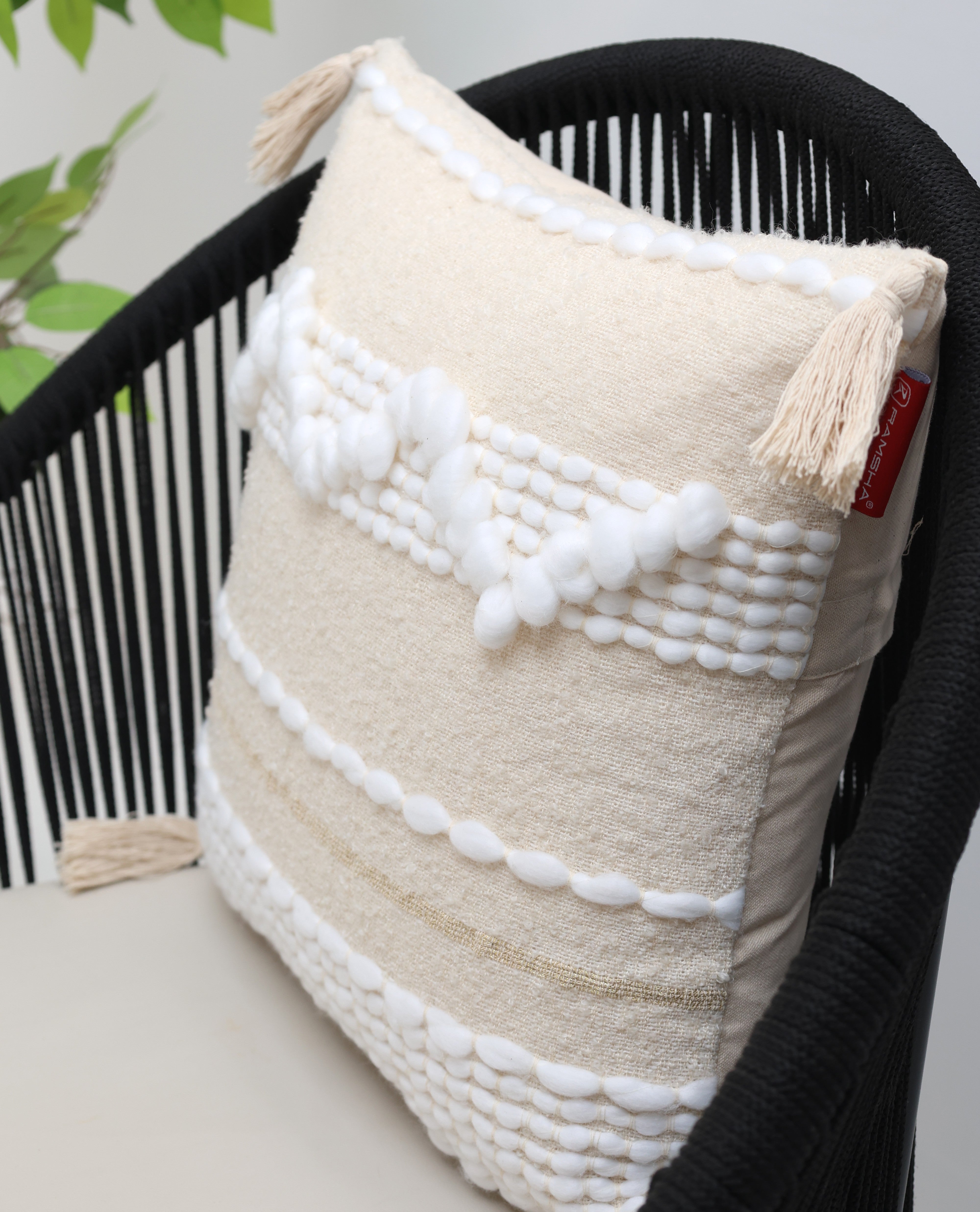 The Cream Color Home Decor Cushion - CS 313 A by Ramsha Home features premium cotton with white textured patterns and tassels, displayed on a black woven chair with a green plant in the background for a stylish décor touch.