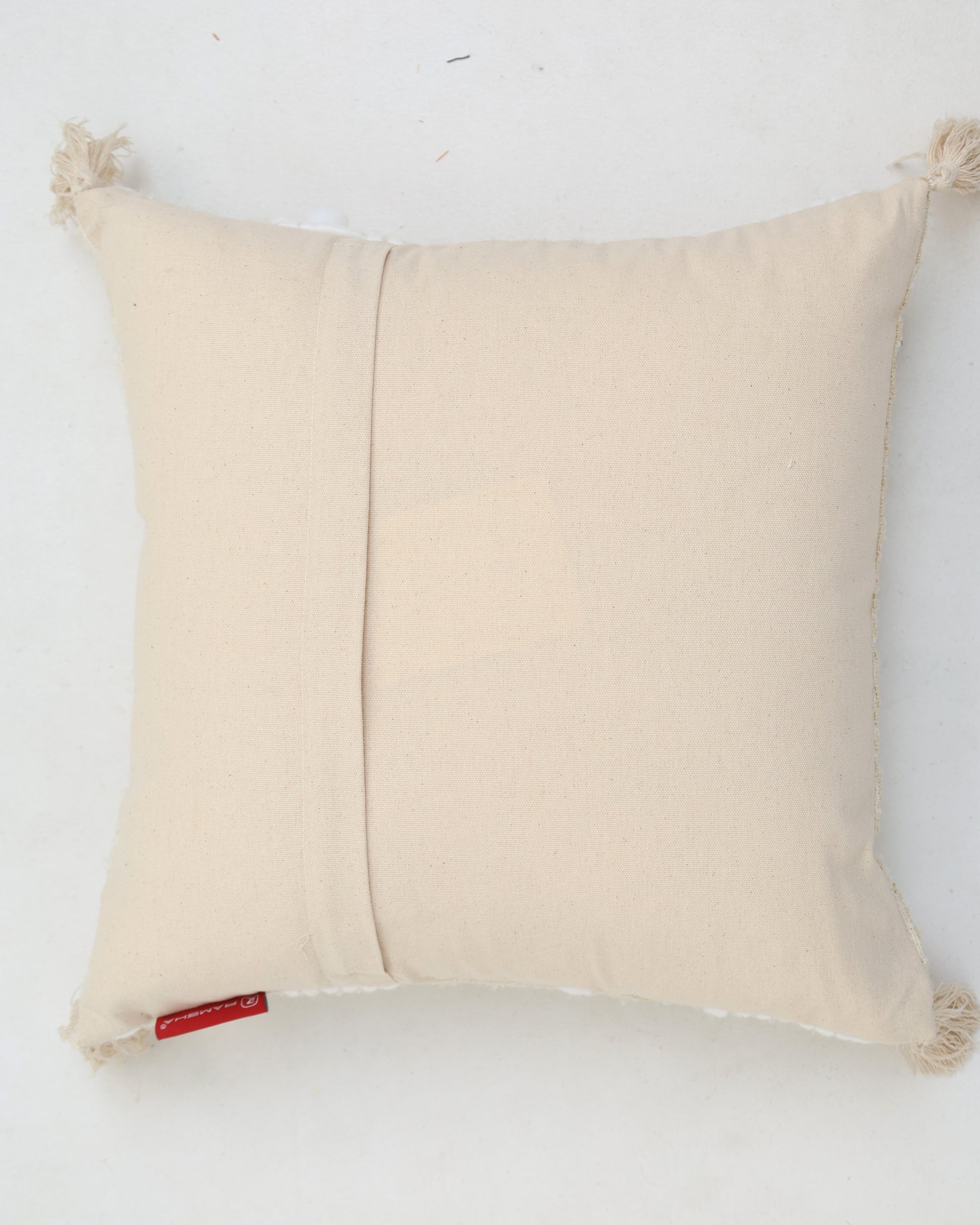 The Cream Color Home Decor Cushion - CS 313 B by Ramsha Home features tassels on each corner, a back pocket, and a red fabric tag. This square cotton-blend cushion is perfect for adding charm to your home décor. Shown on a white background.