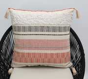 The Ramsha Home Cream Color Home Decor Cushion - CS 314 A features geometric stripes in white, black, beige, and reddish-orange on premium cotton with tassels—ideal for enhancing your décor. Perfect for chairs or sofas.