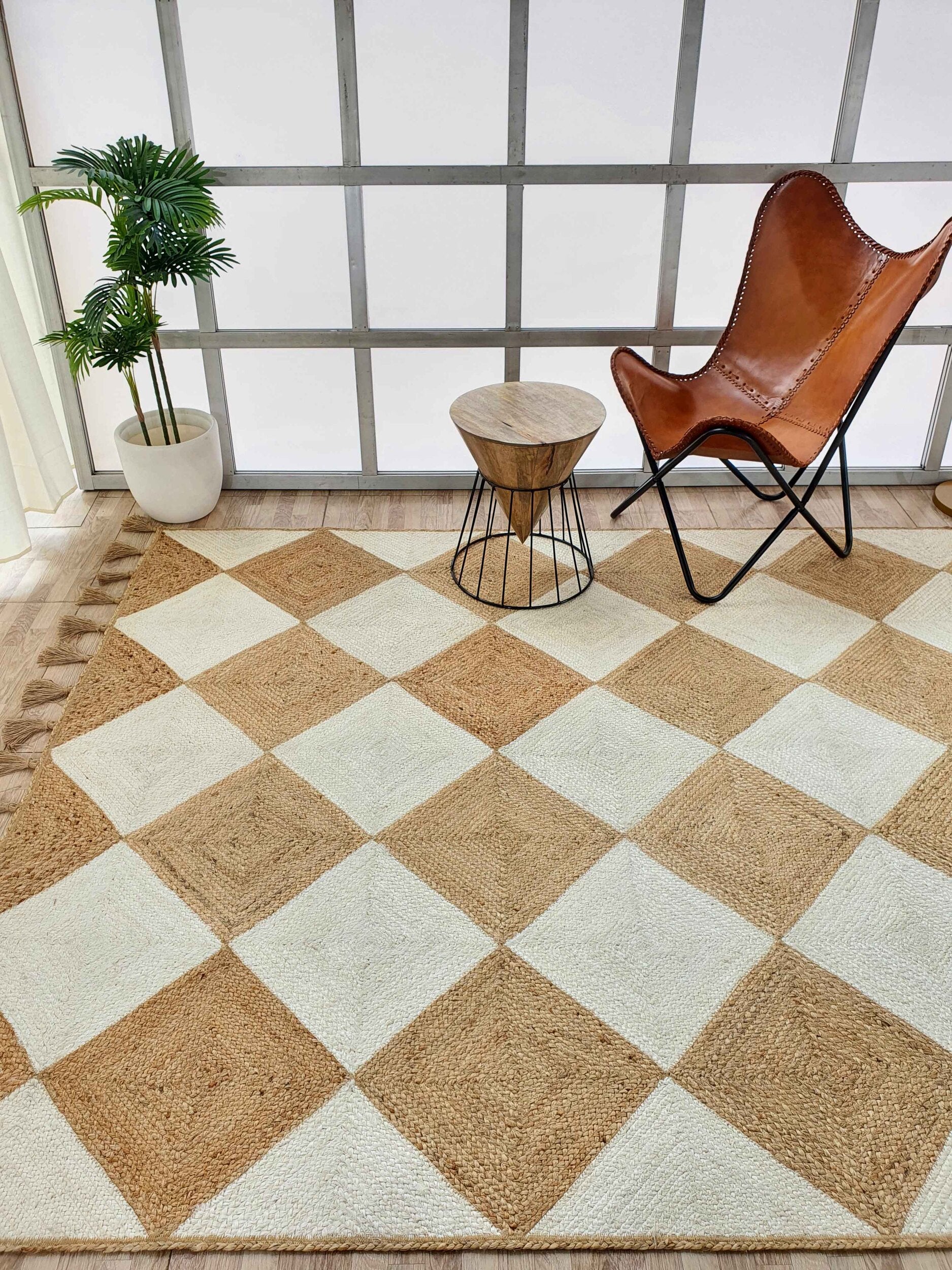 A modern room featuring RAMSHA HOME’s Beautiful Handmade Natural & Bleach Color Jute Braided Rug with Tassel (BR 031), a brown leather butterfly chair, round wood metal-leg side table, potted plant, and large grid-style windows in the background.