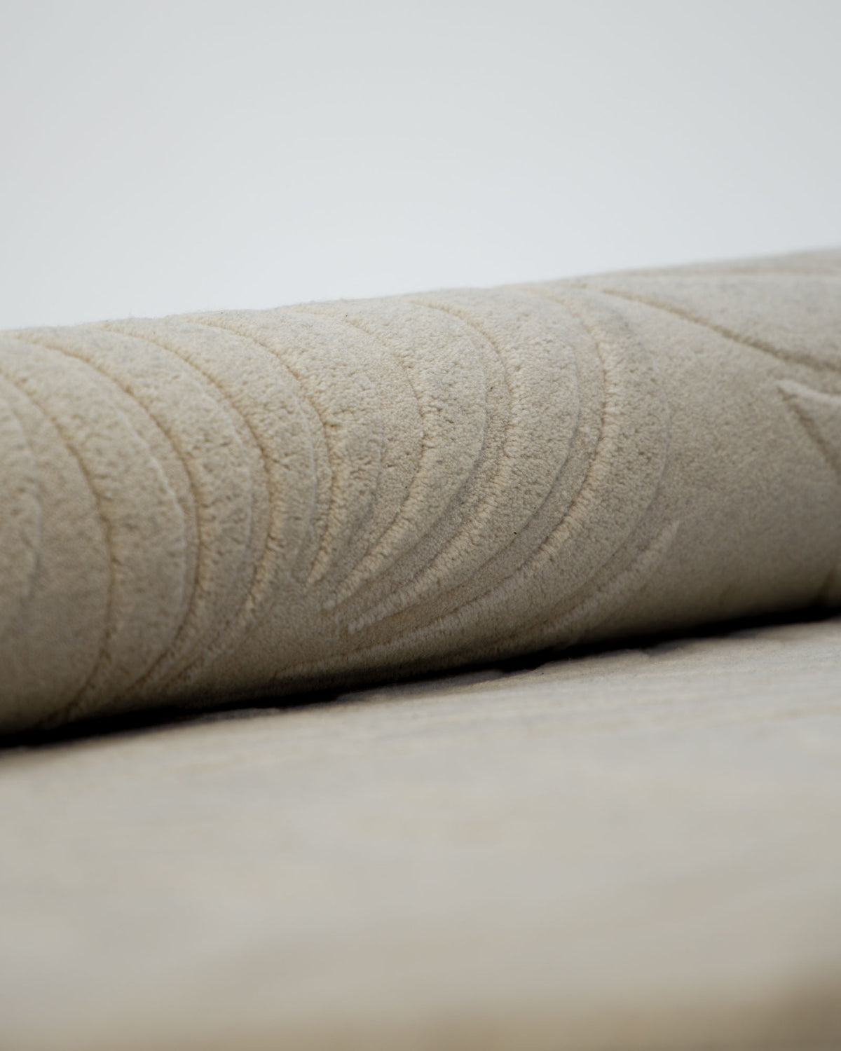 A close-up of a rolled piece of the RAMSHA HOME Premium Off-White Area Carpet - HTW 115 (200x300 cm), showcasing its textured surface and subtle curved patterns, set against a plain, light background.