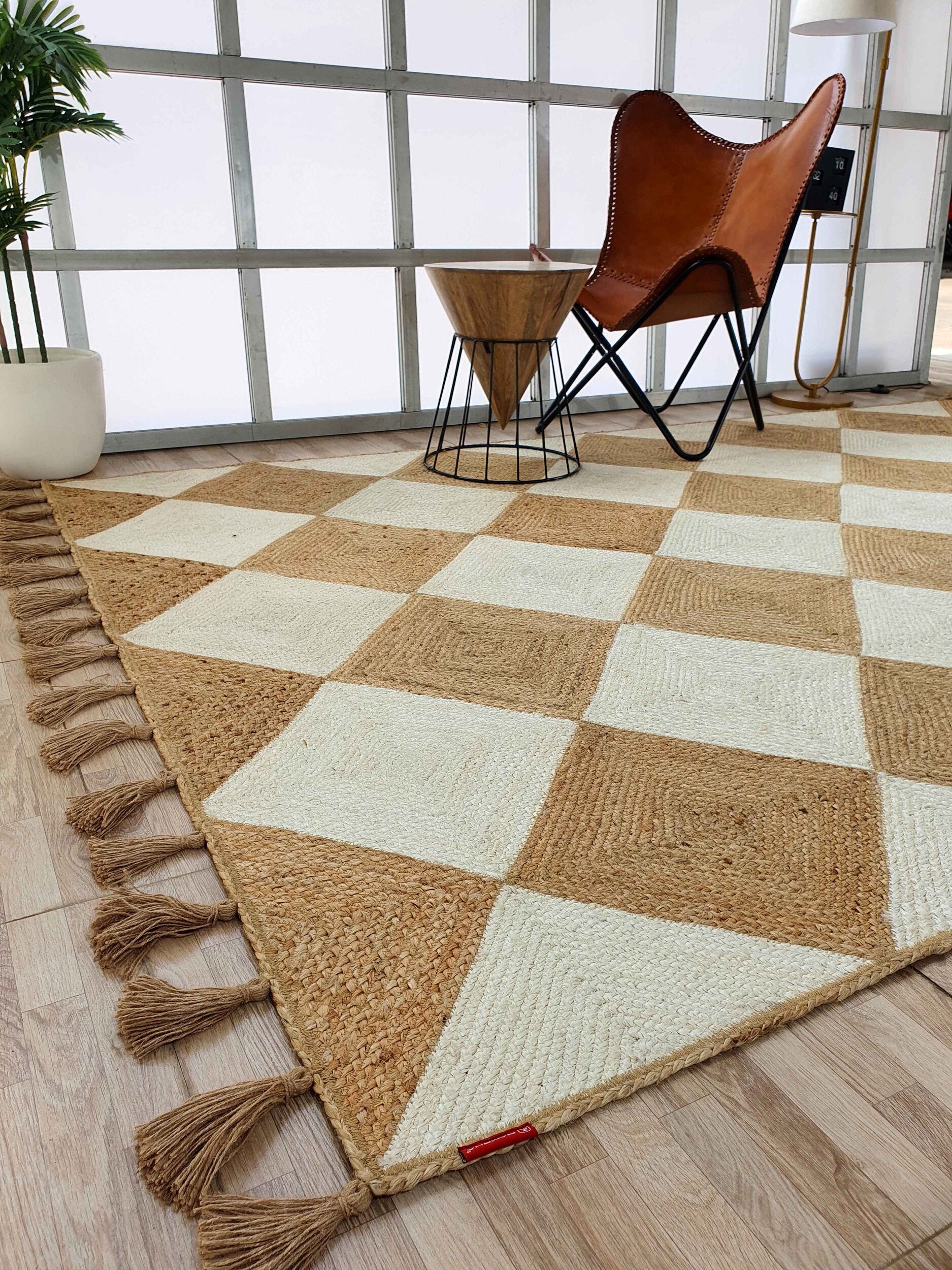 The RAMSHA HOME Beautiful Handmade Natural & Bleach Color Jute Braided Rug with Tassel (BR 031) adds style to a modern room with wood flooring, a leather butterfly chair, geometric wooden side table, potted plant, and grid windows.