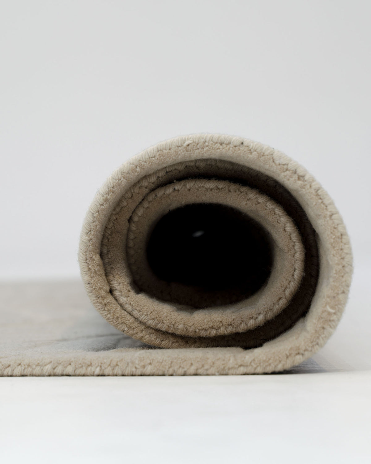 A close-up view of the RAMSHA HOME Premium Off-White Area Carpet - HTW 115 (200x300 cm), rolled up on a light surface with the spiral end facing the camera, set against a plain white background.