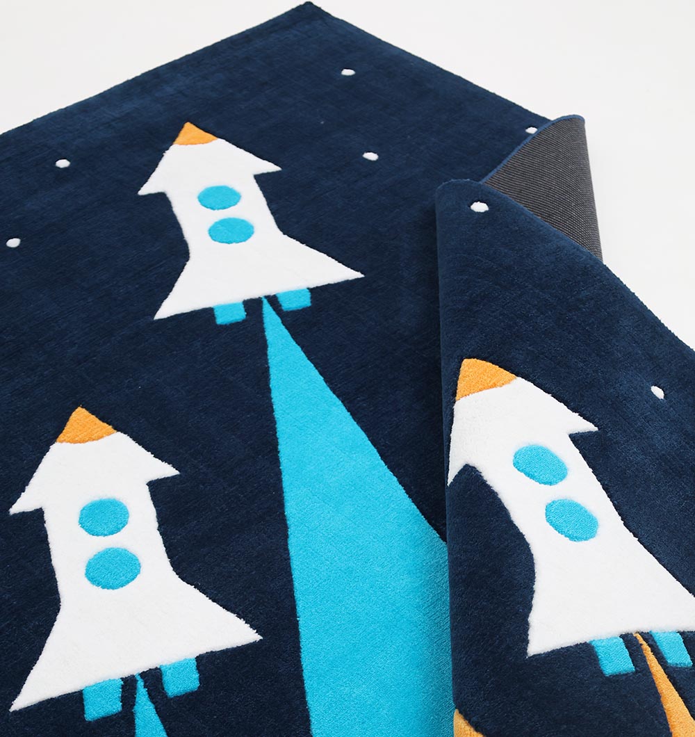 The RAMSHA HOME Kids Space Rocket Polyester Playroom Rug KD-001 (170x240cm) features dark blue tones with white rockets, blue windows, orange tips, and upward blue trails. Part of the playful kids rug is folded over, showing the underside.