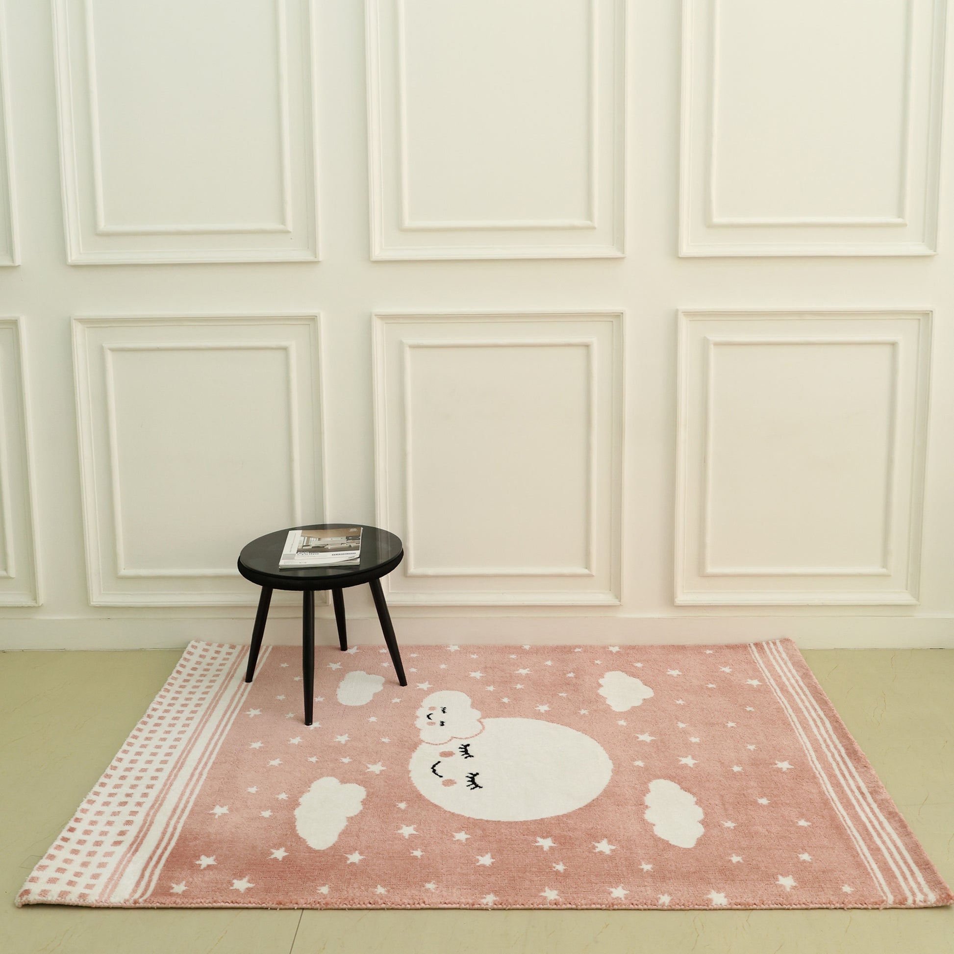 A small round black table with a magazine rests on the RAMSHA HOME Kids Rug Soft Star & Sky Pink Color Nursery Rug (120x180 cm – KD 019), which features a white moon, stars, and clouds, set against a white paneled wall in a minimal room.