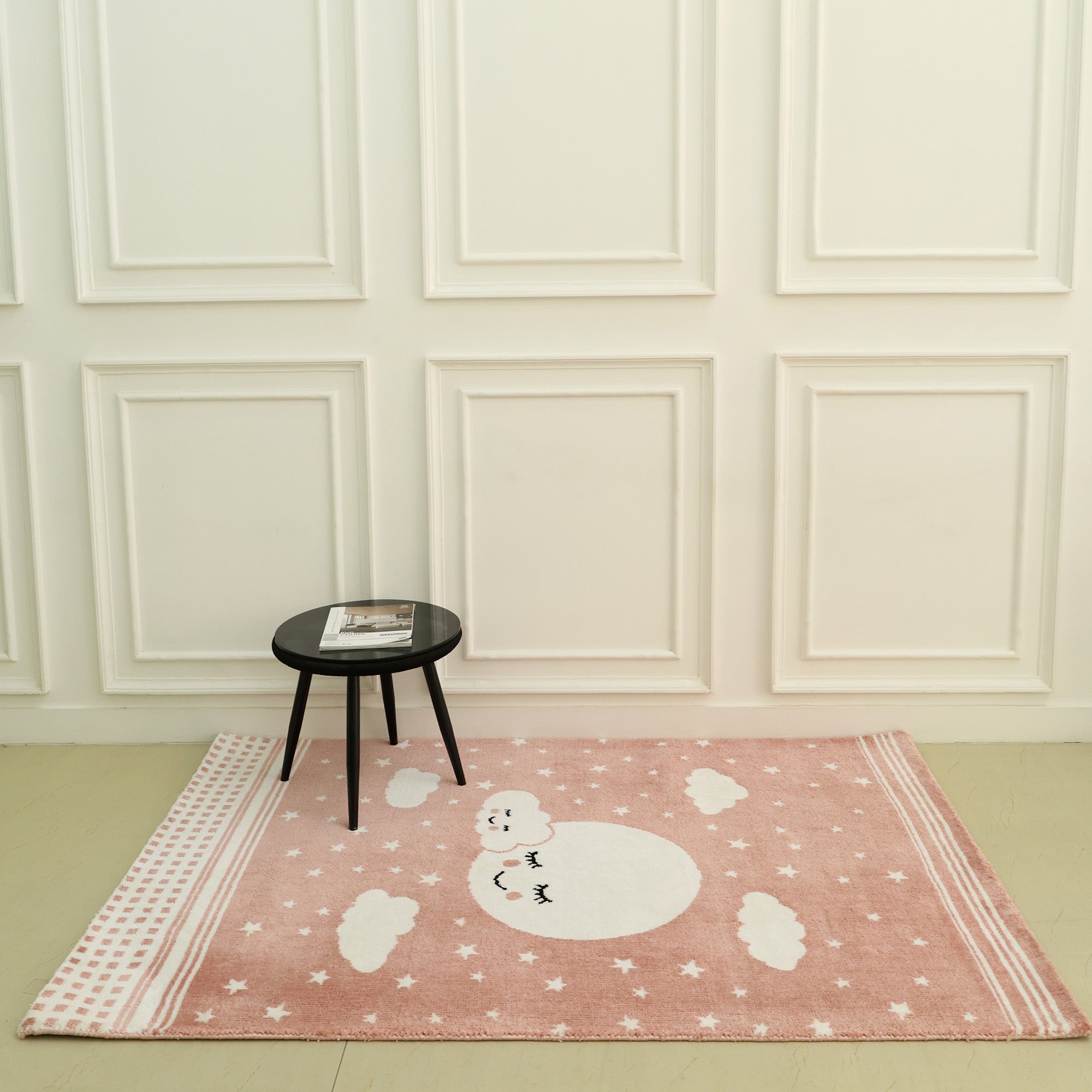 A small round black table with a magazine rests on the RAMSHA HOME Kids Rug Soft Star & Sky Pink Color Nursery Rug (120x180 cm – KD 019), which features a white moon, stars, and clouds, set against a white paneled wall in a minimal room.