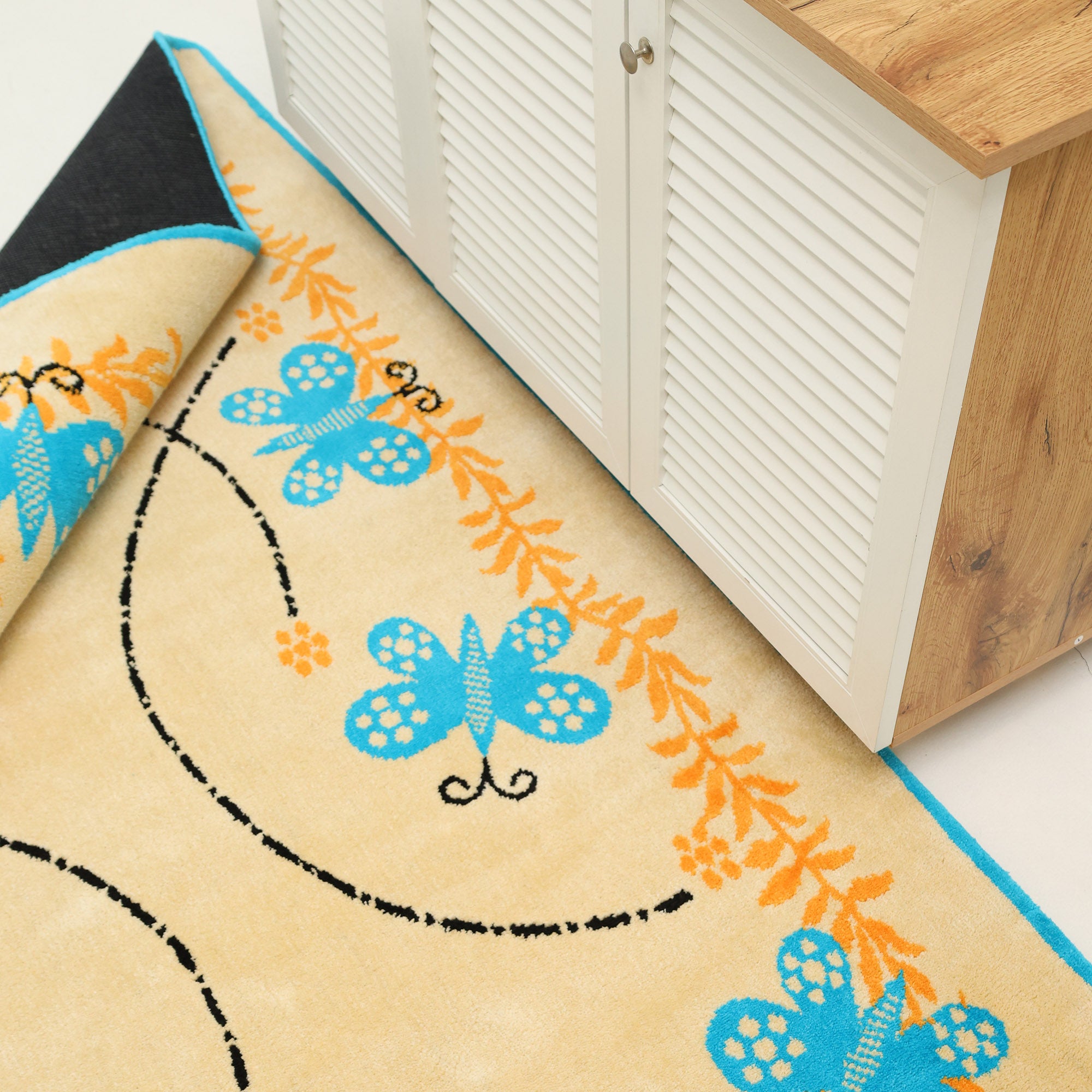 The RAMSHA HOME Kids Rug Soft Multi Color Nursery Rug 120x180 cm – KD 022, featuring blue butterflies and orange floral designs, is partially rolled up beside a white cabinet with louvered doors and a wooden top.