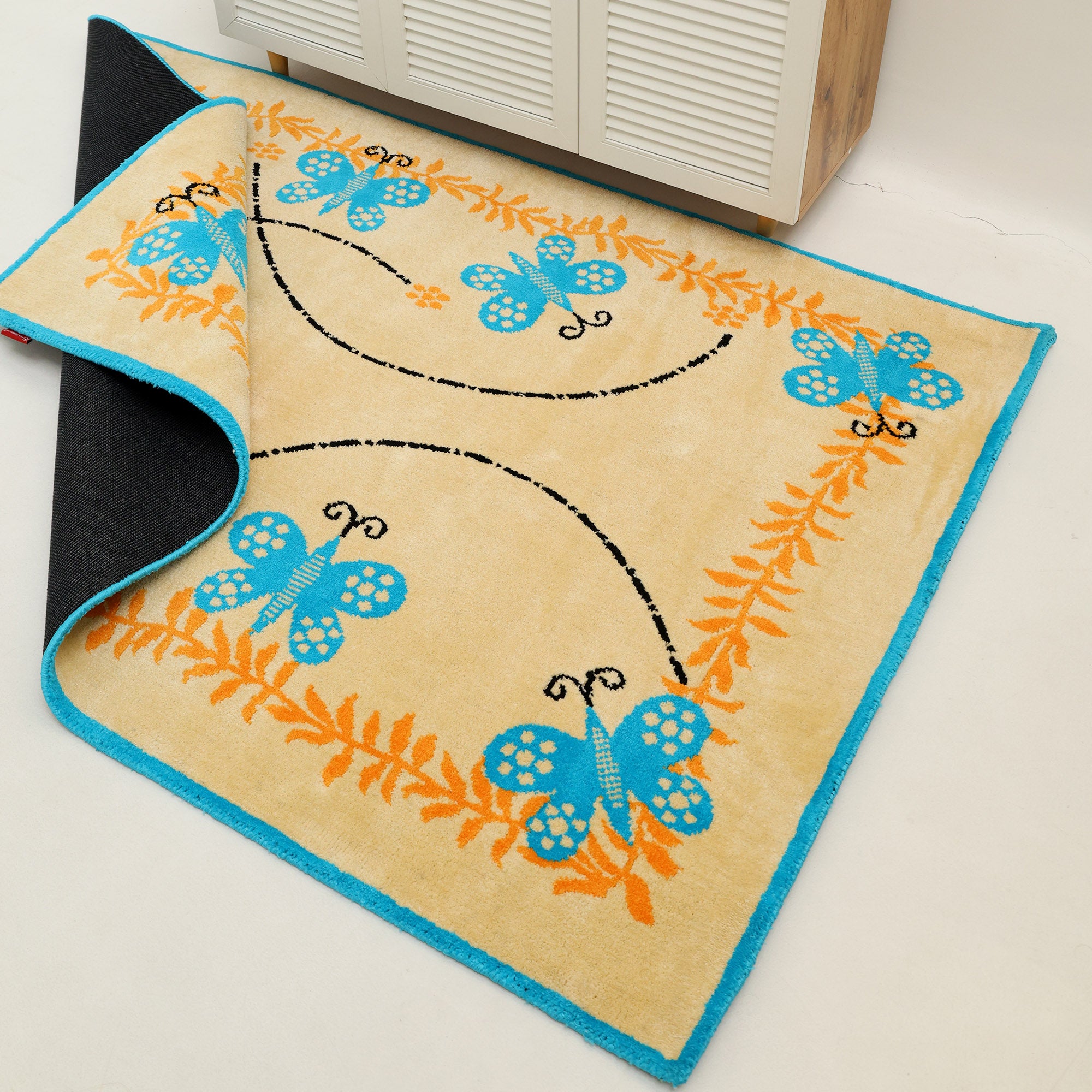 The RAMSHA HOME Kids Rug Soft Multi Color Nursery Rug (120x180 cm – KD 022) features beige, blue butterfly, orange leaf, and black swirl patterns, with a black underside seen at the folded corner. Shown on a light floor near a cabinet.