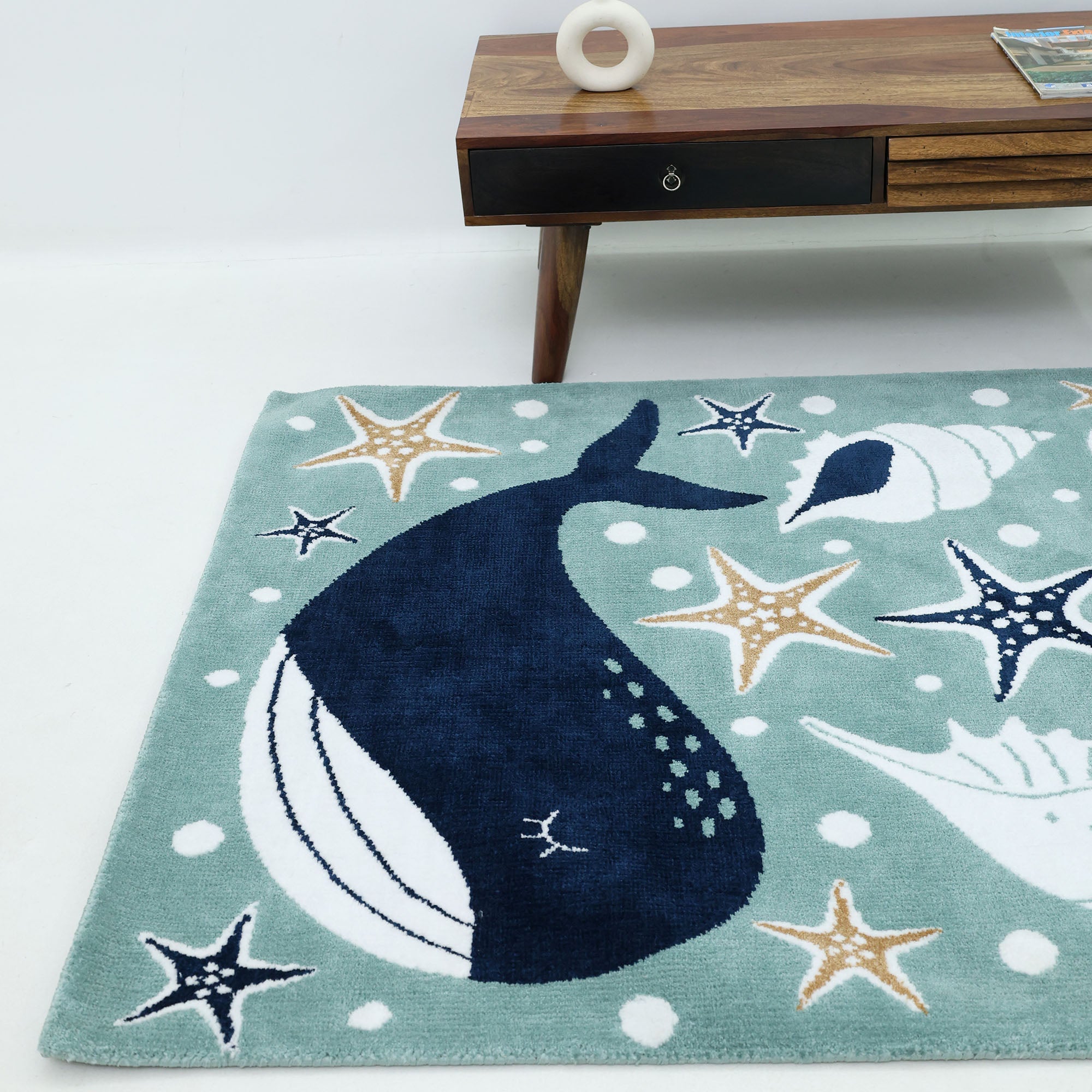 The RAMSHA HOME Kids Rug Soft Whale Character Rug 120x180 cm (KD 017) features a blue whale, sea stars, and gold seashells on a light blue background—perfect for nurseries or play areas in front of tables with decor.