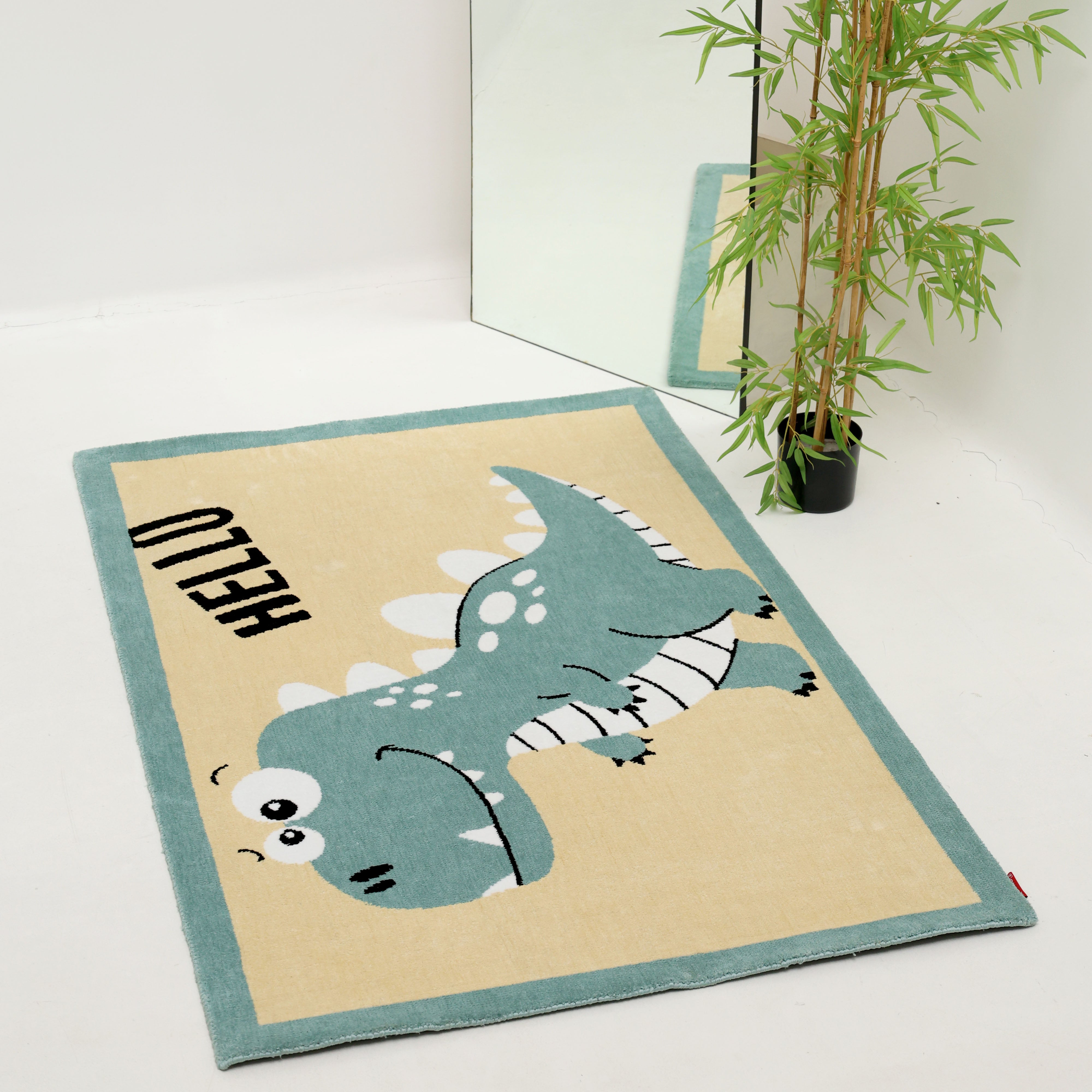 The RAMSHA HOME Kids Rug Soft Dinosaur Character Rug 120x180 cm – KD 018, featuring the word "HELLO," is placed on a white floor near a large mirror and a potted green plant.