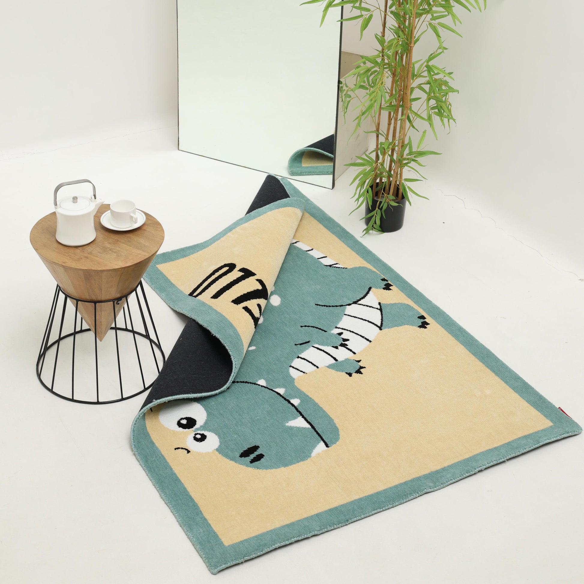 The RAMSHA HOME Kids Rug Soft Dinosaur Character Rug 120x180 cm – KD 018, featuring a playful cartoon dinosaur design, is partly flipped on a white floor beside a tall plant, large mirror, and round table with a teapot and cup.