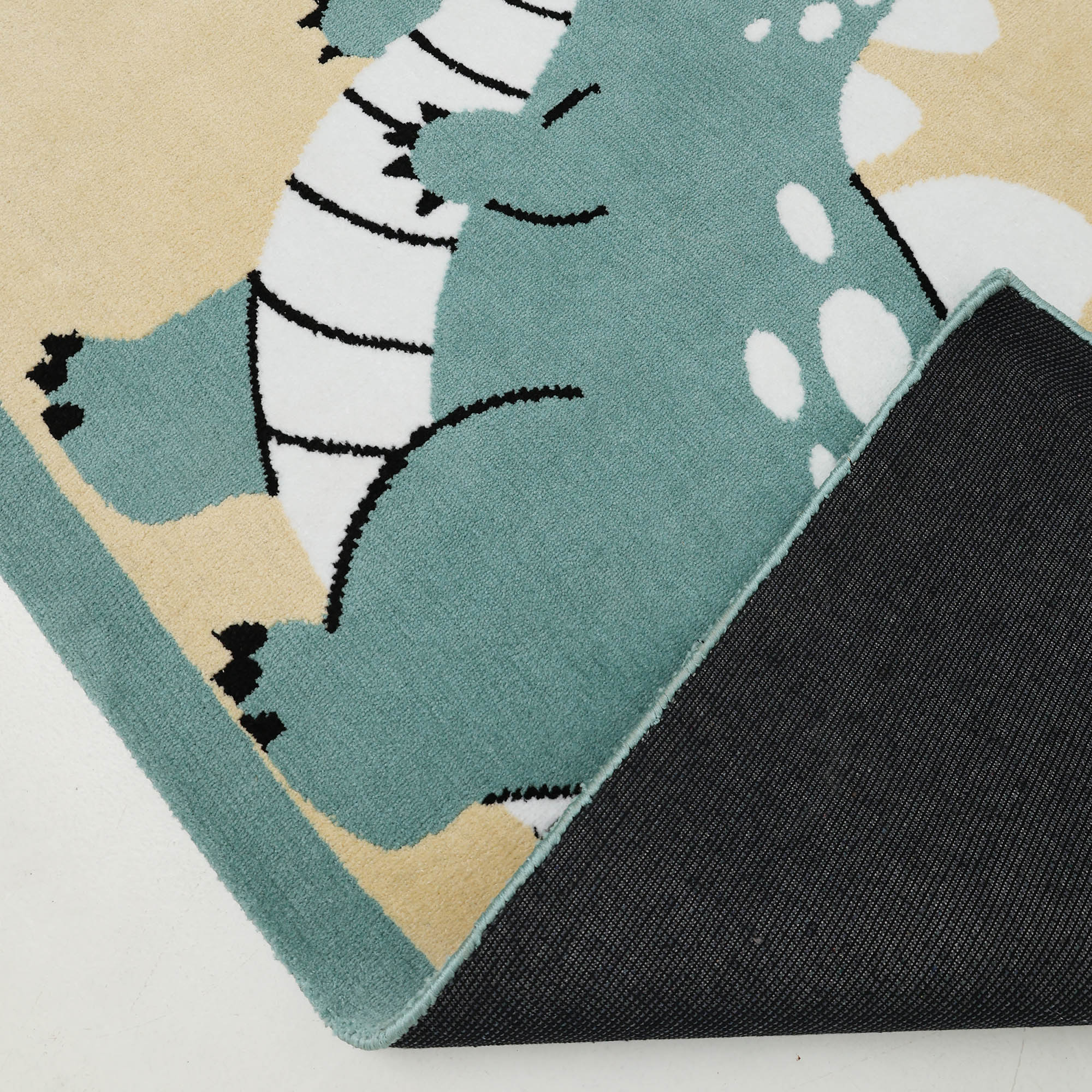 A close-up of the RAMSHA HOME Kids Rug Soft Dinosaur Character Rug (120x180 cm – KD 018), ideal for any kid's room, shows a green cartoon dinosaur with white spots and black stripes and a black textured underside at one lifted corner.