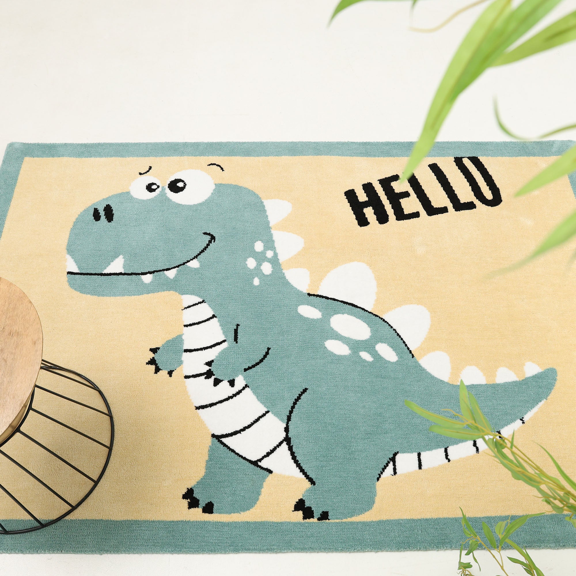 The RAMSHA HOME Kids Rug Soft dinosaur Character Rug 120x180 cm (KD 018) features a cute blue, white-spotted dino and bold "HELLO" on yellow background—perfect playful decor for kids' rooms, with plant and table details at the edges.