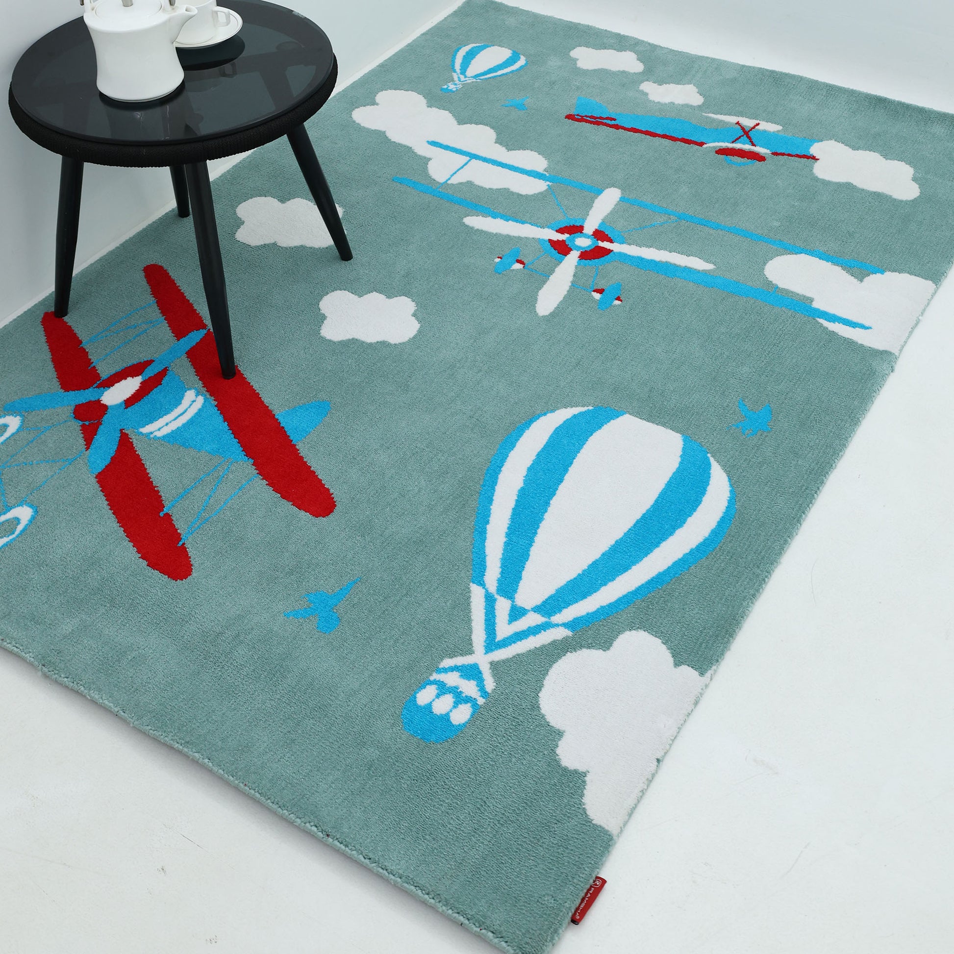 The RAMSHA HOME Kids Rug Soft Multi Color Nursery Rug 120x180 cm – KD 020 is light blue with playful red and blue airplanes, blue and white hot air balloons, and white clouds, shown on a white floor beside a small black table with a teapot and cups.