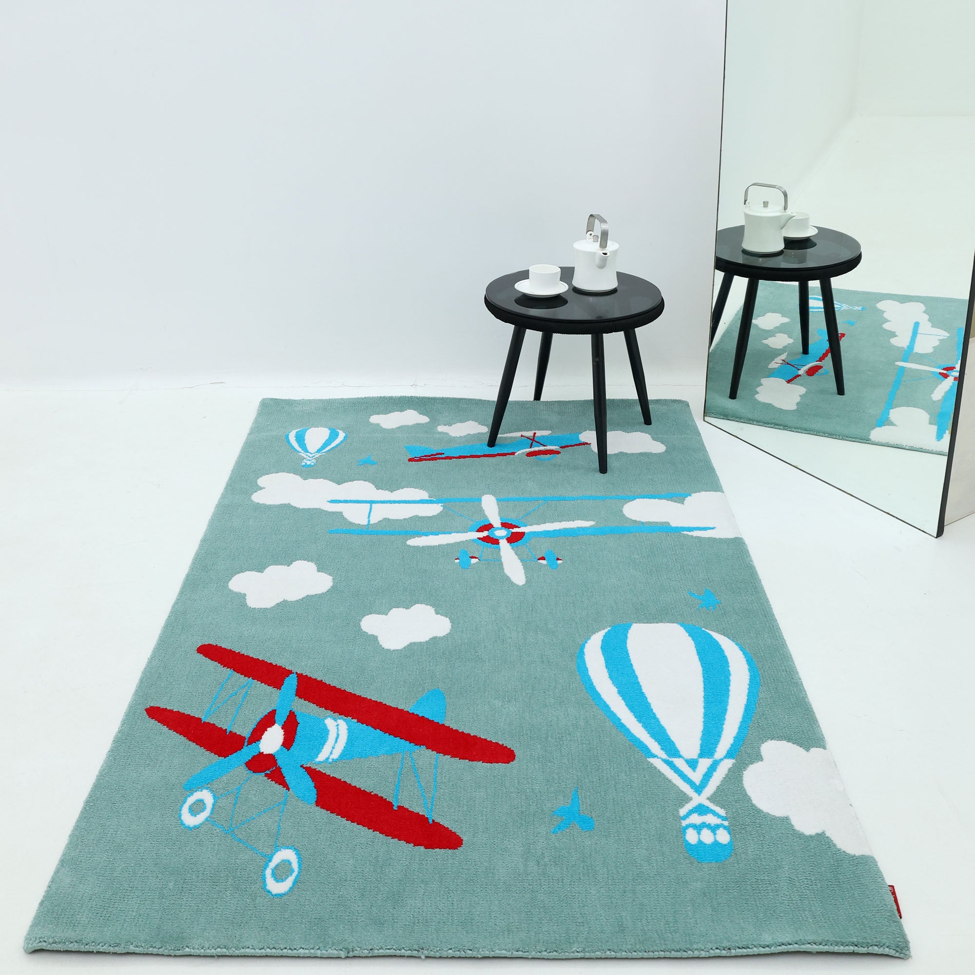 The RAMSHA HOME Kids Rug Soft Multi Color Nursery Rug 120x180 cm (KD 020), with playful airplane, balloon, and cloud designs, is on a white floor. Nearby, a small black table with a white teapot and cups sits beside a large mirror.