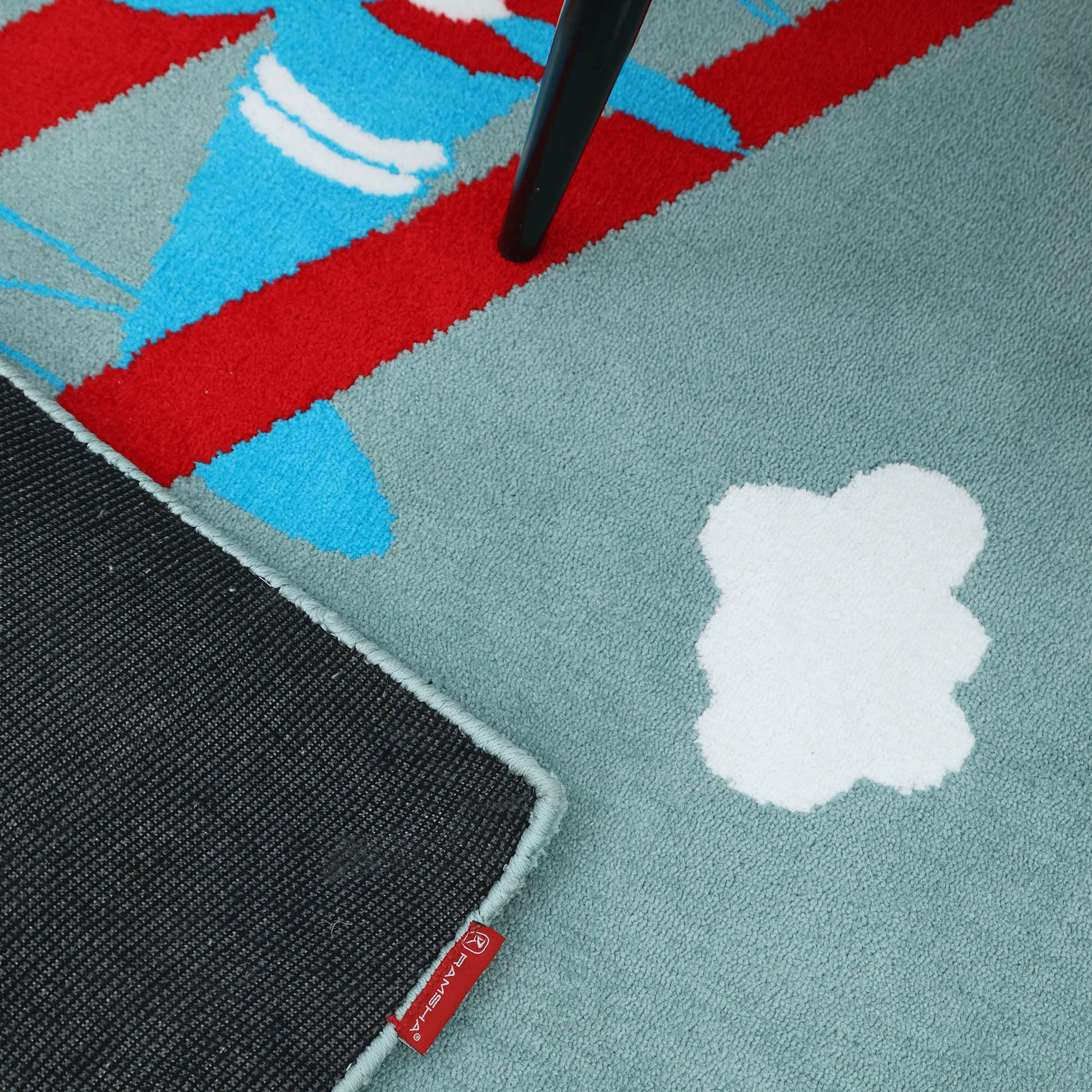 A close-up of the RAMSHA HOME Kids Rug Soft Multi Color Nursery Rug (120x180 cm, KD 020) shows white cloud shapes, red and blue abstract patterns, a black rug layered on top with a red fabric tag, and a chair leg in the corner.