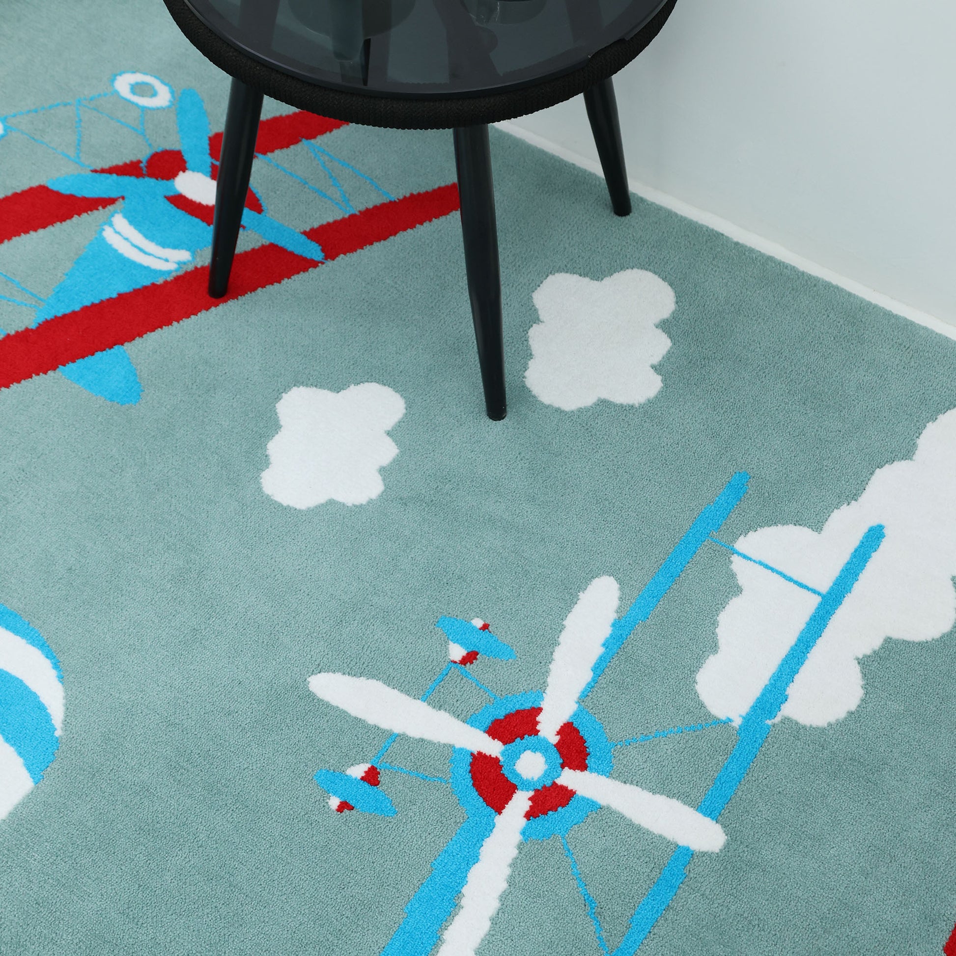 The RAMSHA HOME Kids Rug Soft Multi Color Nursery Rug 120x180 cm – KD 020 features red, white, and blue vintage airplane and cloud illustrations on a light blue background, with a black round glass table leg visible in one corner.
