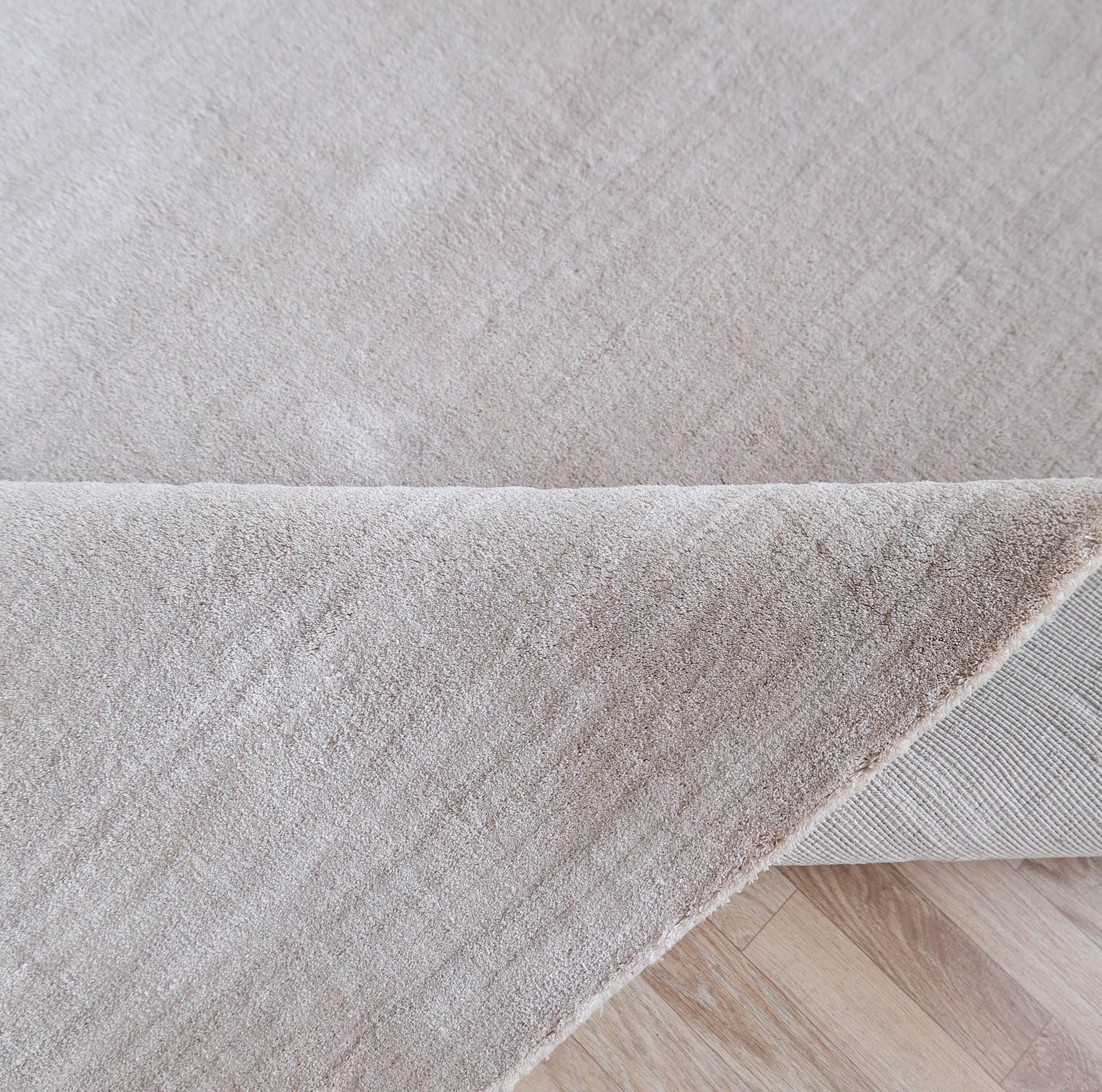 A close-up of the RAMSHA HOME Handmade Plain Beige Area Color Rug 300x400 cm (LKH-047), partially folded on a wooden floor, highlights its soft textured surface and sturdy backing.