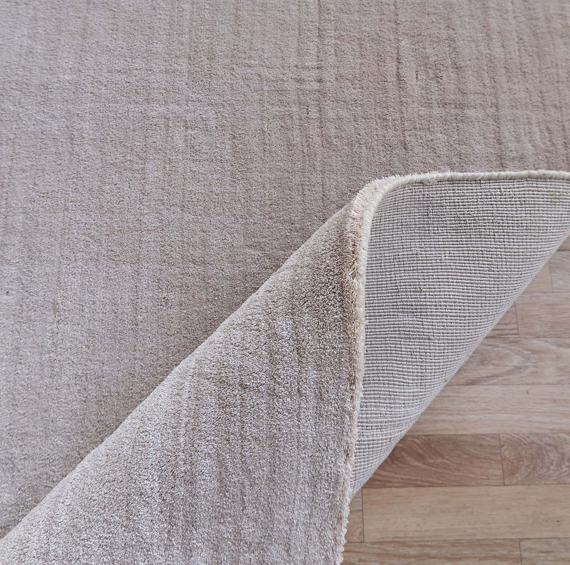 The RAMSHA HOME Handmade Plain beige Area Color Rug (300x400 cm, LKH-047) rests on a light wooden floor, with one cream-toned corner lifted to reveal its handwoven texture beneath.