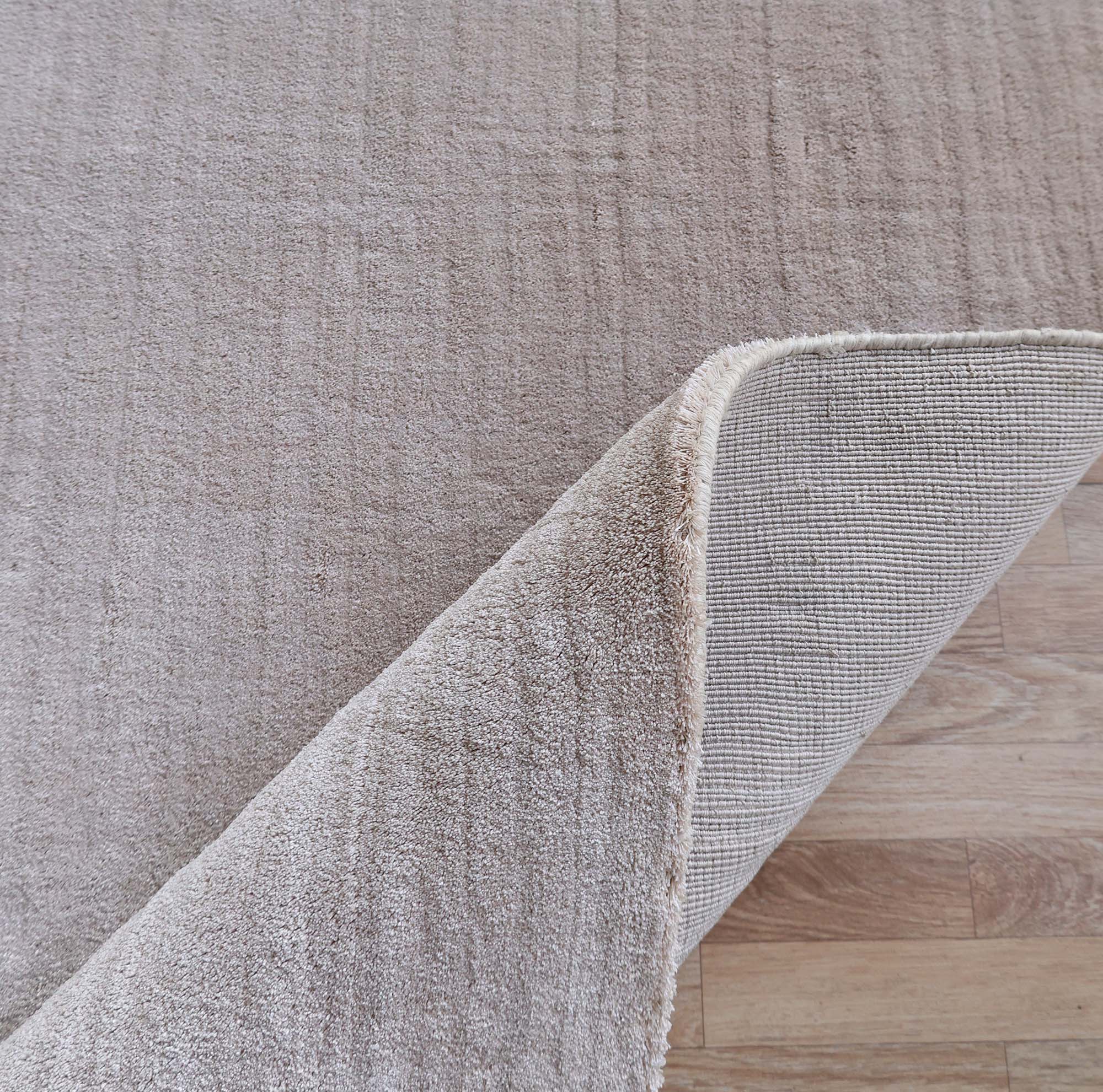 The RAMSHA HOME Handmade Plain beige Area Color Rug (300x400 cm, LKH-047) rests on a light wooden floor, with one cream-toned corner lifted to reveal its handwoven texture beneath.