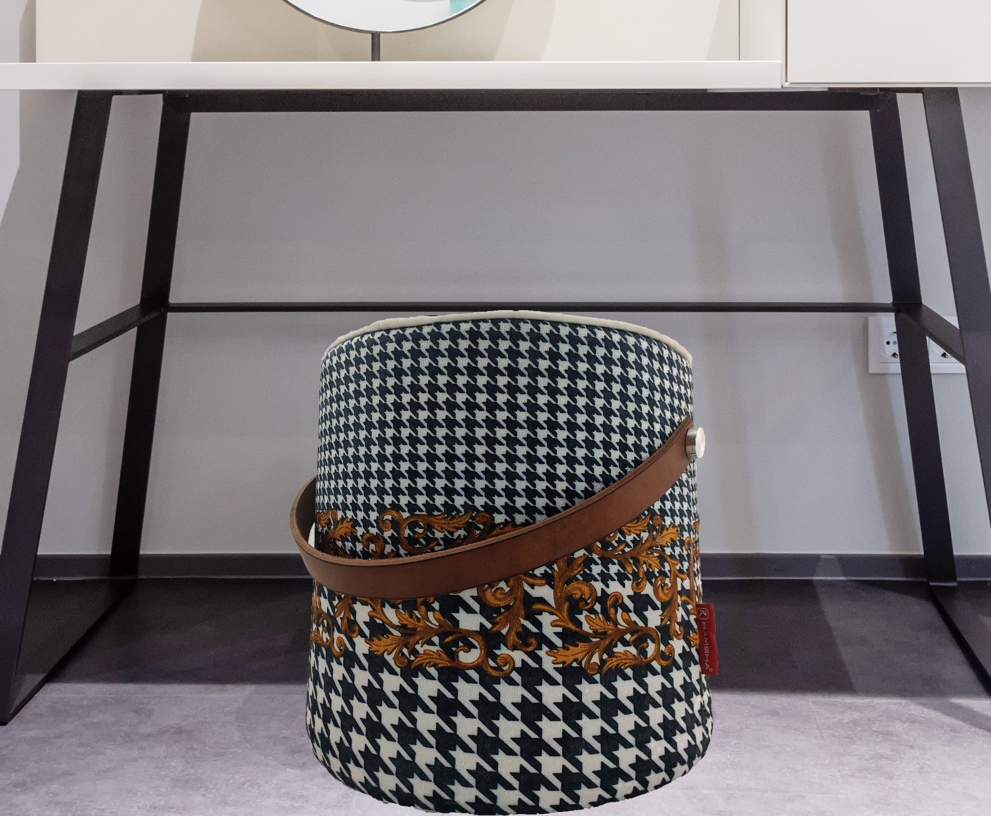The RAMSHA HOME Leather Printed Pedestal Pouf With Leather Handle PF-025, featuring a black-and-white houndstooth print, gold floral accents, and a brown leather handle, sits stylishly under a modern desk—an elegant home decor accent.