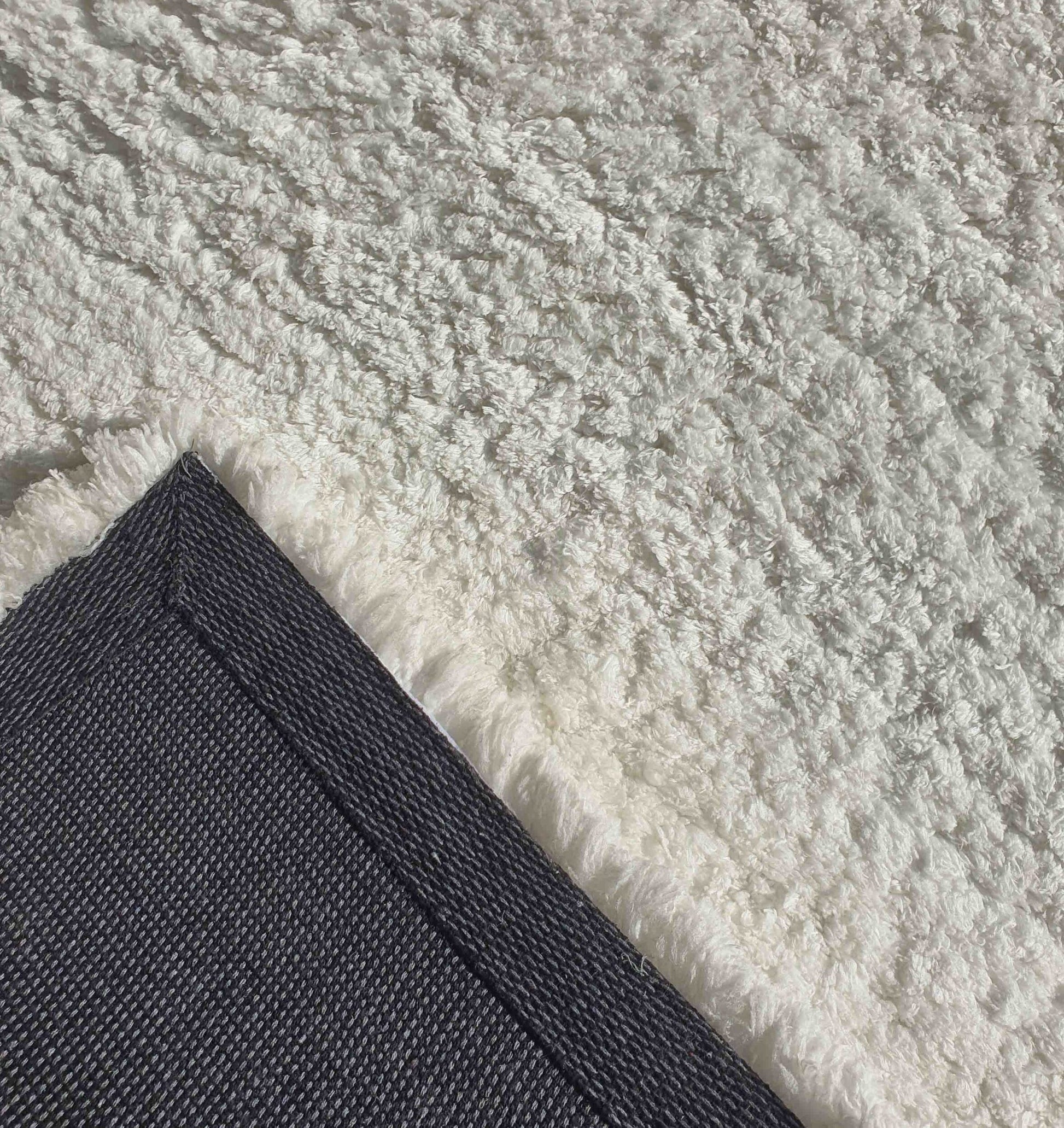 A close-up of the RAMSHA HOME Soft White Rectangle Bedroom Rug Shaggy Rug - SG 06 shows its pebbled shag texture, with one corner flipped to reveal the black woven backing and white fringe, while bright sunlight highlights its soft surface.